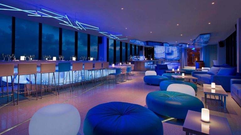 Altitude Rooftop Bar toạ lạc tại tầng cao nhất được bao quanh bởi các khung cửa kính trong suốt, ghế bar cao và ghế classic.